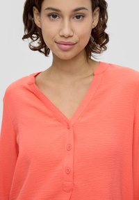 Korallfarbene V-Ausschnitt-Bluse mit strukturiertem Finish, Knopfleiste und kleinem Kragen. Lange Ärmel und weicher Stoff.