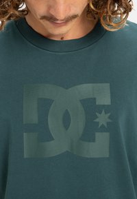 Türkisfarbiges Baumwoll-T-Shirt mit einem großen, tonalen DC-Logo-Graphic auf der Vorderseite, ergänzt durch einen Stern-Akzent und einem Rundhalsausschnitt. Glatte Textur.