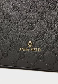 Sac à main noir texturé avec des motifs circulaires en relief, arborant un logo doré "ANNA FIELD" au centre. Forme rectangulaire, design élégant.