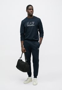 Mladý muž oblečený v tmavě modré teplákové soupravě EA7 Emporio Armani a bílých teniskách, držící černou kabelku přes rameno, stojící před jednobarevným pozadím.