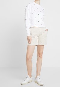 Vitt kort svetttröja med färgglada logotryck, parat med beige shorts och vita sneakers. Slät tyg, avslappnad passform, inga tydliga mönster.