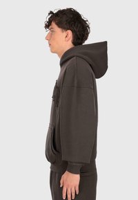Giovane uomo in profilo che indossa un ampio felpa nera con cappuccio, dotata di tasca frontale, e pantaloni abbinati neri, su uno sfondo chiaro e semplice.