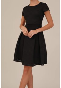 Femme portant une robe noire texturée à manches courtes et longueur genou, avec des bagues et un bracelet en or, debout devant un fond beige uni.