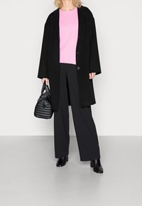 Schwarzer übergroßer Mantel, rosa strukturiertes Oberteil, schwarze weit geschnittene Hose und schwarze Stiefel mit Absatz. Accessoire: kleine schwarze gesteppte Handtasche.