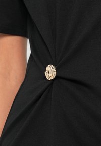 Μαύρο μπλουζάκι με στριφογυριστό σχέδιο μπροστά, που διαθέτει ένα διακοσμητικό χρυσό κουμπί στο κέντρο. Λείο ύφασμα.