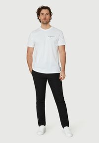 Vit bomulls t-shirt med en liten svart logo på vänster bröst, i kombination med svarta byxor och vita sneakers, mot en enfärgad bakgrund.