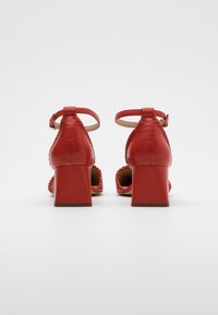 Chaussures à talon bloc en cuir rouge avec un design de bride tressée et une boucle à la cheville. Texture lisse et talon de hauteur moyenne.