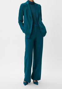 Türkisfarbener Damen-Blazer und weite Hose Set, aus glattem Stoff gefertigt. Bemerkenswerte Details sind ein tailliertes Rollkragenoberteil und spitze Absätze.
