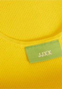 Tissu côtelé jaune avec une étiquette rectangulaire verte cousue près du bord affichant les lettres blanches "JJXX" centrées.