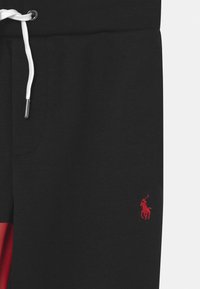 Černé tepláky s hladkou texturou, vybavené bílou šňůrkou a malým červeným logem. Detail boční kapsy a design v barevných blocích v červené barvě.