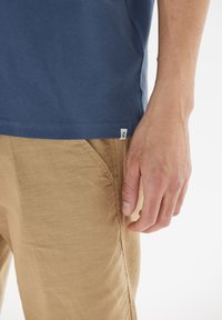 Blå bomulls t-shirt med korta ärmar, logotagg i sidans söm; kombinerad med ljusbruna strukturerade byxor som framhäver en figurnära siluett och handposition.