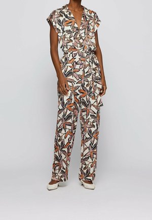 Jumpsuit floral en crème avec des motifs de feuilles orange et noir, présentant un design enveloppant, des manches courtes et une ceinture à nouer. Coupe ajustée.