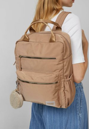 Femme portant un sac à dos en nylon beige avec deux poches avant zippées et un porte-clés en fourrure ronde, vêtue d'un haut blanc et d'un jean bleu.