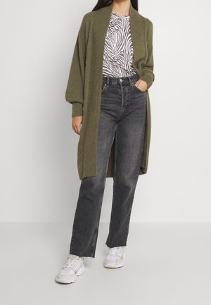 Woman wearing long olive green cardigan over zebra print top, high-waisted faded black jeans, and white sneakers standing against plain background.