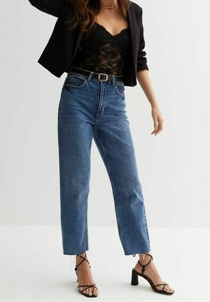 Kvinde iført sort blonder top, kort sort blazer, blå højtaljede jeans med bælte og sorte blokketsandal, står mod hvid baggrund.