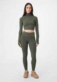 Nainen seisoo yllään oliivinvihreä istuva urheilupaita, samanväriset legginsit ja beigeet lenkkarit tasaisella valkoisella taustalla.