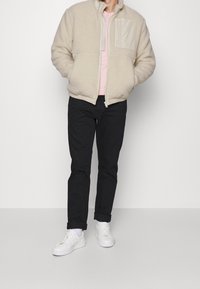 Ljusbeige, fluffig zip-up jacka med ficka fram; bärs över en ljusrosa skjorta, kombinerad med svarta byxor och vita sneakers.