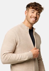 Cardigan de cremallera color beige claro, tejido texturizado, con cuello y puños acanalados, usado sobre una camisa negra, que muestra un ajuste ceñido.
