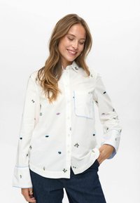 Glunderende vrouw met lang haar, gekleed in een witte blouse met kleine kleurrijke geborduurde ontwerpen en donkerblauwe spijkerbroek, met haar hand in haar zak.