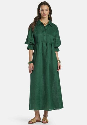 Femme portant une longue robe boutonnée vert foncé avec des manches bouffantes, des bijoux en or et des sandales tissées à enfiler, se tenant devant un fond blanc.