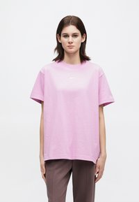 Noor naine seisab, seljas lõdvavus roosa Nike t-särk ja pruuni värvi püksid, heleda valge tausta ees.