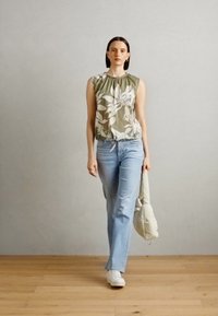 Ljusgrön blommig topp med rynkad halsringning, i kombination med ljusblå jeans. Modell med vita sneakers som håller en ljusbeige väska.