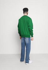 Sweatshirt verde in cotone con profili a coste; indossato con pantaloni in denim blu sbiadito; design semplice; il modello è di spalle rispetto alla telecamera.