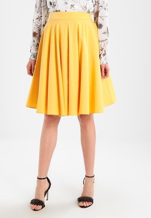 Nainen, joka pukeutuu kirkkaan keltaiseen laskostettuun hameeseen, kukalliseen pitkähihaiseen puseroon ja mustiin korkeakorkoisiin sandaaleihin, joissa on koristeellisia piikkejä.