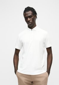 Jeune homme aux cheveux tressés portant un polo blanc à manches courtes avec une fermeture éclair à l'avant, debout les mains dans les poches d'un pantalon beige.