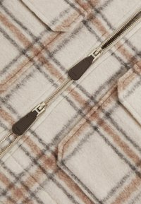 Gros plan sur un tissu à carreaux beige et marron avec une double fermeture éclair et une poche plaquée, montrant un matériau en laine texturé.
