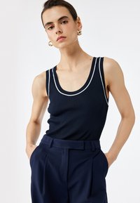 Marineblaues geripptes Tanktop mit weißem Besatz, breiten Trägern und Rundhalsausschnitt, kombiniert mit hoch taillierten marineblauen Hosen. Glatte Textur sichtbar.