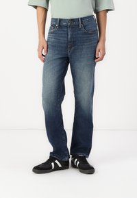 Modré denimové džíny se středně vysokým pasem a rovným střihem, s kontrastním prošíváním a klasickými kapsami. Nosené s černými teniskami.