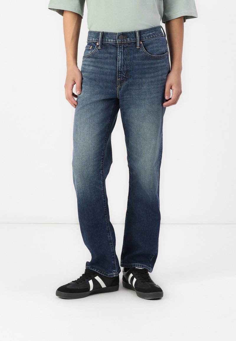 Modré denimové džíny se středně vysokým pasem a rovným střihem, s kontrastním prošíváním a klasickými kapsami. Nosené s černými teniskami.