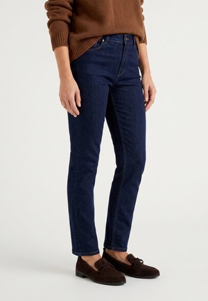 Person trägt dunkelblaue Slim-Fit-Jeans, braune Loafers und einen braunen Pullover vor einem einfarbigen hellen Hintergrund.