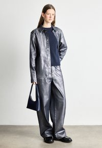 Jaqueta de denim metálica com botões, combinada com calças largas a condizer. Suéter azul marinho por baixo. A modelo segura uma pequena bolsa azul marinho.