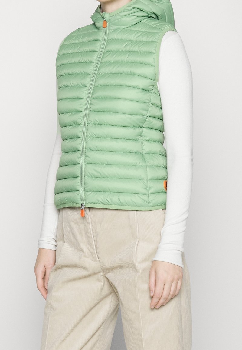 Ljust grön, zip-up pufferväst med huva, med horisontell quiltad design och en orange dragkedjehandtag. Bärs över en vit långärmad skjorta.