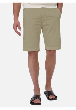 Homme portant un short beige mi-cuisses et des sandales noires à enfiler, debout, les mains détendues, jambes visibles de la taille aux pieds.