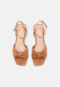 Bruine suède sandalen met een vierkante neus, knoopstrik detail aan de voorkant en een verstelbaar enkelbandje. Zachte textuur en minimalistisch ontwerp.