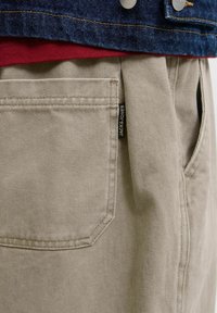 Μπεζ βαμβακερό παντελόνι με πίσω τσέπη και μαύρη ετικέτα "JACK & JONES", φορεμένο με κόκκινο πουκάμισο και μπλε denim μπουφάν με μεταλλικά κουμπιά.