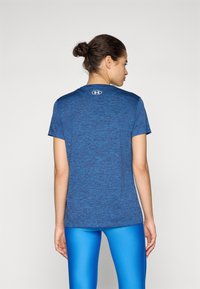 Camiseta deportiva azul de manga corta, con tejido texturizado, cuello redondo y logo en la parte trasera. Combinada con leggings azules, vista desde atrás.