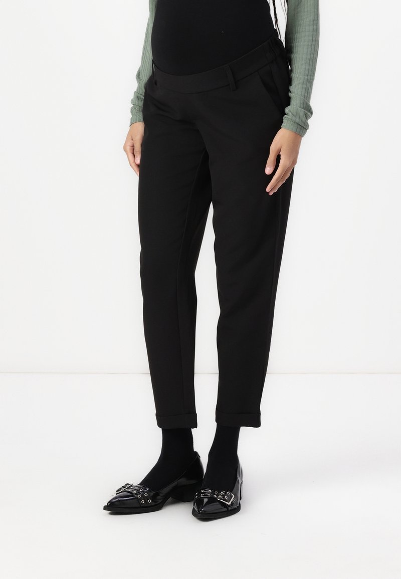 Partie basse du corps d'une personne portant un pantalon noir raccourci, des collants noirs, des mocassins noirs brillants avec détails de boucle et œillets, et un haut vert à manches longues côtelé.