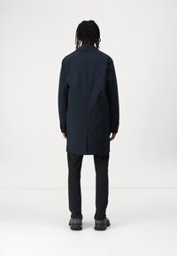 Langer navyblauer Mantel aus glattem Stoff mit geradem Schnitt, mit Kragen und knöpfbaren Manschetten. Getragen über dunklen Hosen und Schuhen.
