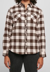 Femme portant une chemise à carreaux marron et blanche avec deux poches poitrine et un pantalon noir, debout devant un fond uni.