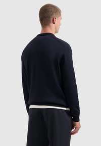 Man in een zwart longsleeve trui en zwarte broek, gezien van achteren tegen een effen lichte achtergrond.