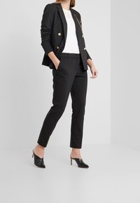 Blazer nero con bottoni dorati, top bianco e pantaloni neri aderenti. Silhouette snella con tacchi alti neri a punta. Design minimalista, texture liscia.