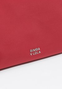Czerwona torebka wykonana z tkaniny o gładkiej fakturze, z wytłoczonym na przodzie srebrnym logo "BIMBA Y LOLA". Prosty, nowoczesny design.