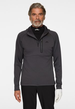 Pull à fermeture éclair gris foncé fabriqué dans un tissu doux avec un col haut, comportant une petite poche poitrine et un logo noir sur le côté gauche.