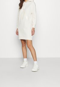 Krämfärgad hooded klänning med långa ärmar, framficka och ribbat fåll, tillsammans med vita sportskor och ankelsockor.