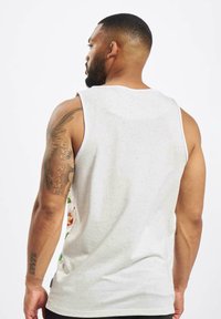 Hvid ærmeløs tanktop med spottet tekstur, blomsterdesign på siderne og rund halsudskæring. Synlige tatoveringer på venstre arm.