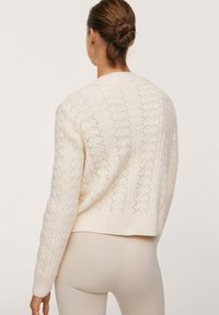 Kobieta z brązowymi włosami zwiniętymi w kok, ubrana w kremowy sweter w warkocze i beżowe dopasowane spodnie, pokazana od tyłu na jednolitym tle.